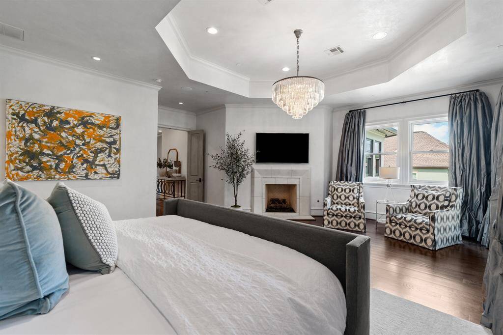 This additional view of the primary suite showcases a serene sitting area framed by oversized windows, a cozy fireplace beneath a sparkling crystal chandelier, and elegant tray ceilings that elevate the sense of luxury and retreat.