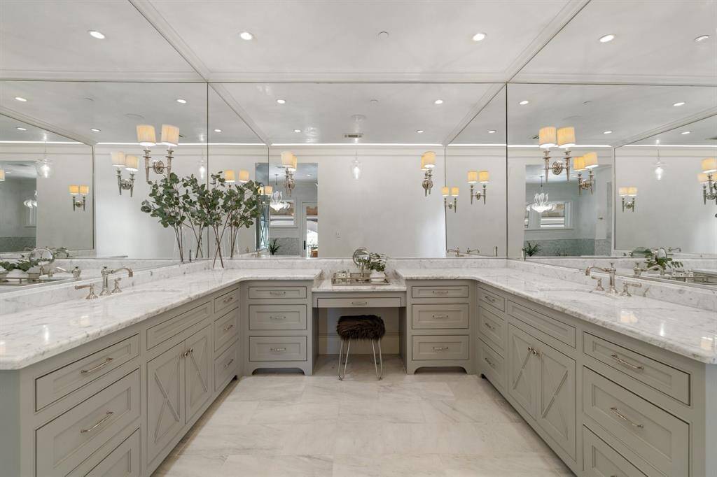 This exquisite primary bathroom features dual vanities with elegant marble countertops, custom cabinetry in a soft taupe hue, and a dedicated makeup station, all illuminated by warm sconces and recessed lighting. Expansive wall-to-wall mirrors enhance the sense of space and luxury, creating a spa-like ambiance that is both functional and refined.