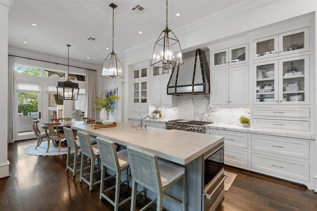 This stunning chef’s kitchen blends timeless elegance with high-performance design, featuring sleek white cabinetry, dramatic marble countertops and backsplash, and a statement black metal range hood. A spacious island with seating for four anchors the space, illuminated by stylish pendant lighting and seamlessly connecting to the light-filled breakfast area and outdoor living beyond.