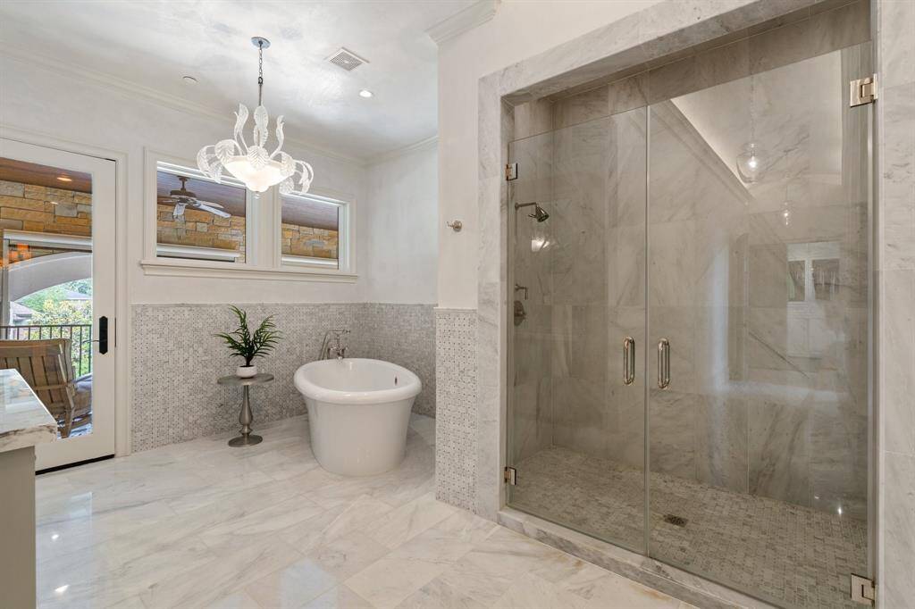 This luxurious primary bathroom is a serene sanctuary, featuring gleaming marble floors, a spa-like walk-in shower with dual showerheads, and a sculptural freestanding soaking tub beneath an artful glass chandelier. French doors open to a private covered terrace, blending indoor elegance with outdoor tranquility.