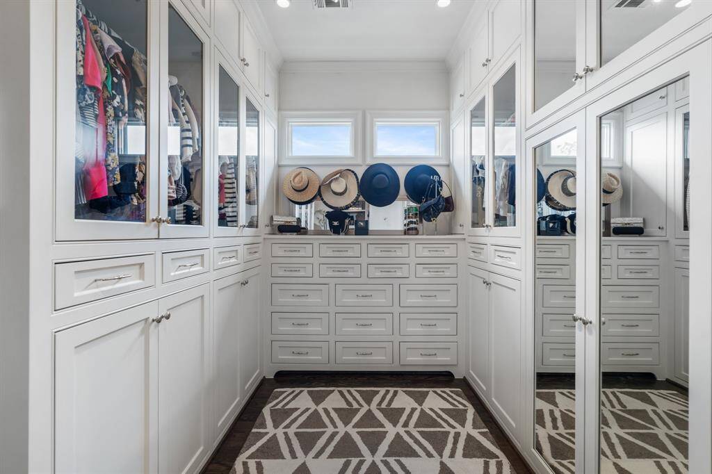 The expansive primary closet showcases custom built-ins with glass-front cabinetry, abundant drawer storage, and chic display space all beautifully illuminated by natural light from dual transom windows.