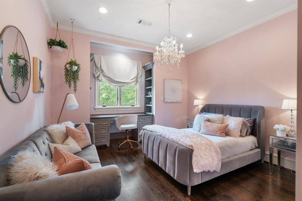 This chic secondary bedroom features soft blush-toned walls, a crystal chandelier, and a tufted velvet bed that create an atmosphere of comfort and elegance. A built-in desk nook with shelving offers a functional workspace, while the cozy sitting area adds a perfect spot for relaxation.