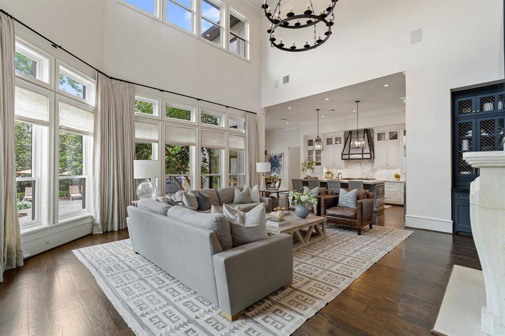 Bathed in natural light from a soaring wall of windows, this elegant living room offers a grand yet inviting atmosphere with its double-height ceilings, statement chandelier, and seamless flow into the gourmet kitchen. Rich hardwood floors, layered textures, and sophisticated furnishings create a refined space perfect for both relaxed family living and stylish entertaining.