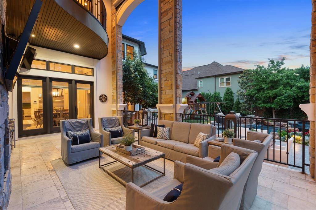 This inviting covered patio offers a sophisticated outdoor living experience, framed by grand stone columns and arched architecture. Overlooking a resort-style pool and lush landscaping, the space is ideal for relaxed gatherings or evening lounging under ambient lighting.