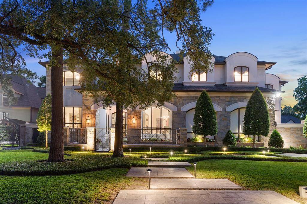 This twilight exterior captures the grandeur of a stately custom estate, showcasing impeccable architecture framed by manicured landscaping and dramatic evening lighting. Soaring arched windows, a stone façade with stucco accents, and elegant wrought iron details exude timeless sophistication, while a stone path flanked by uplighting draws the eye toward the warmly illuminated entryway, inviting guests into a world of refined luxury.
