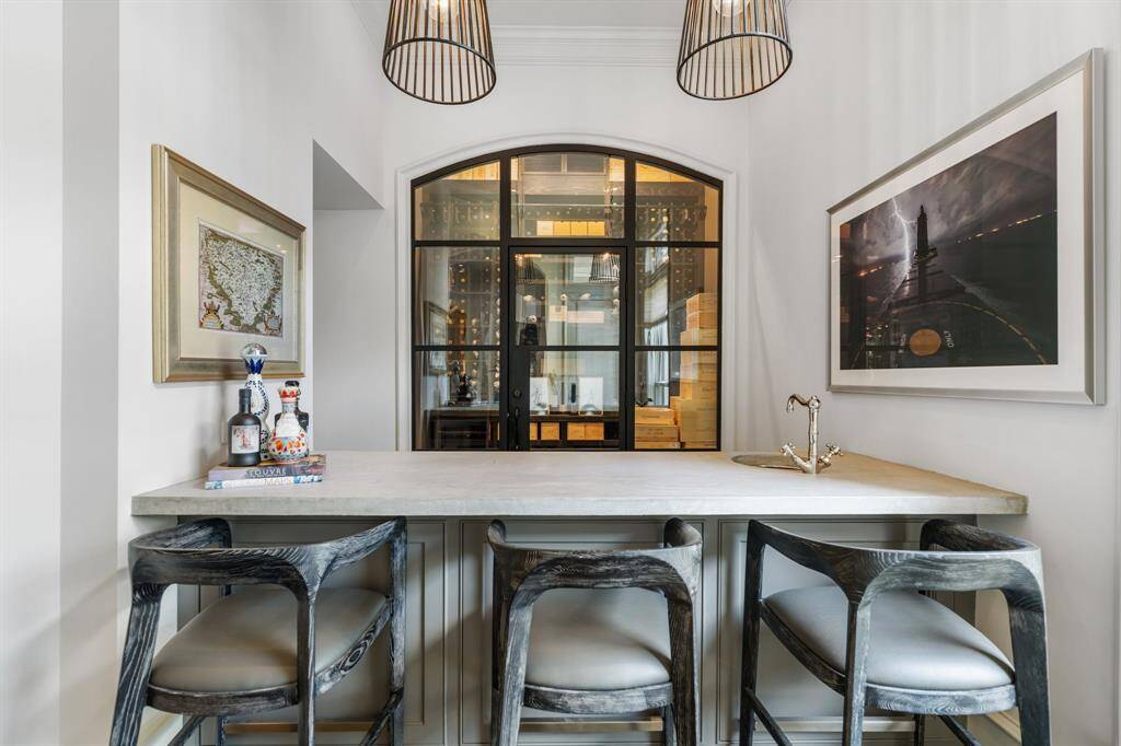 This stylish bar area is both functional and inviting, featuring a sleek concrete countertop with integrated sink, designer pendant lighting, and artisan bar stools for a curated touch. A striking arched glass wall reveals a climate-controlled wine cellar beyond, creating a dramatic focal point that elevates the space into an entertainer’s dream.