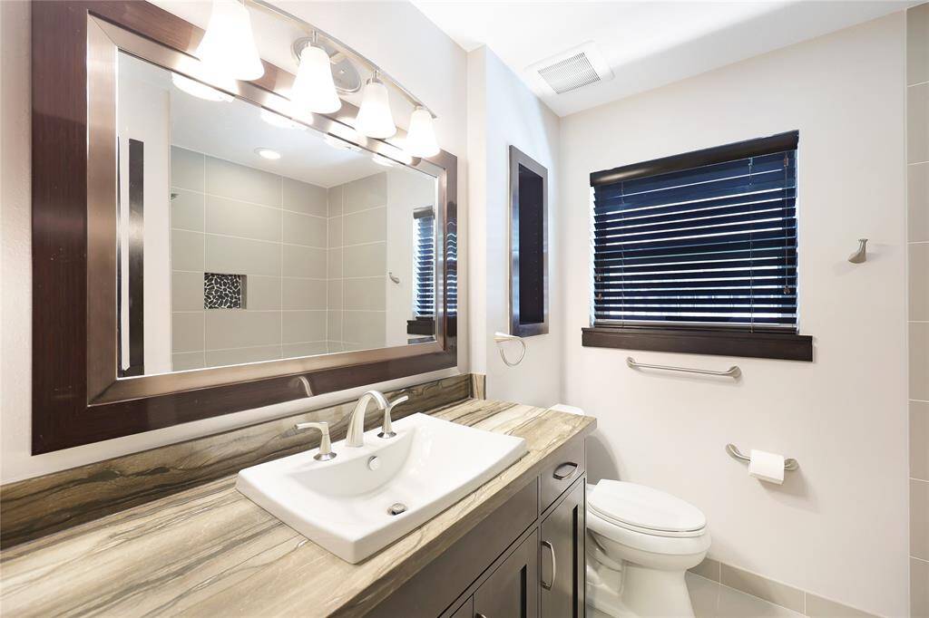 Guest bathroom with a stylish vanity, large mirror, and sleek lighting. Features a tiled shower area and a window with blinds for natural light.