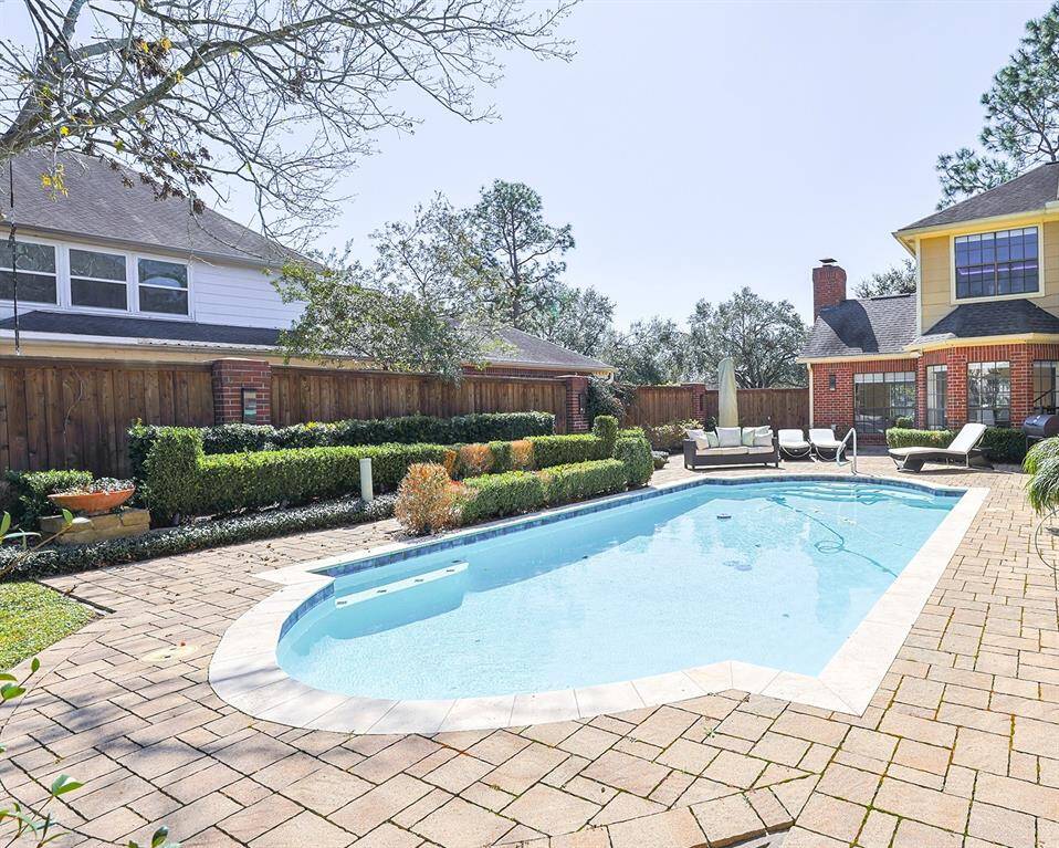 A private backyard retreat featuring a sparkling swimming pool surrounded by lush greenery and manicured hedges. The spacious stone-paved patio is perfect for lounging, and the backyard is equipped with a mosquito misting system for added comfort.