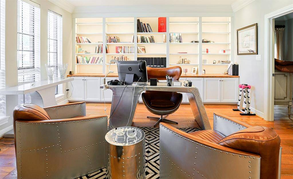 A sleek and modern office space with built-in bookshelves, providing ample storage and display options. Large windows allow for plenty of natural light, while the contemporary lighting fixture adds an elegant touch. The stylish metallic and leather furniture gives the space a sophisticated, industrial-chic vibe.