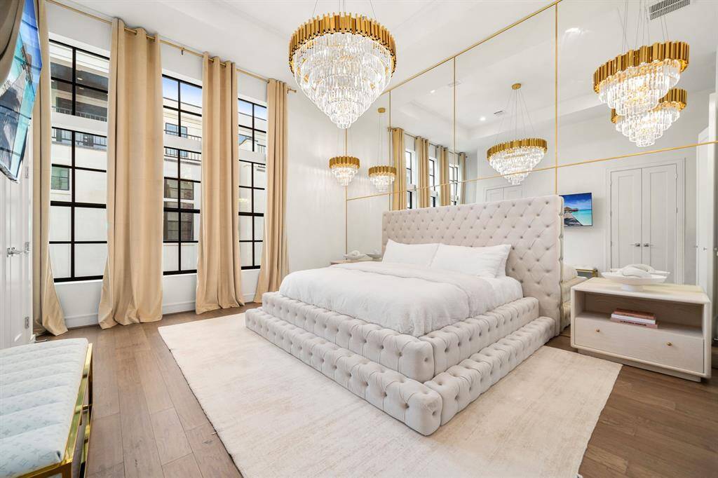 This elegant primary bedroom features a luxurious tufted bed, high ceilings, and large windows with flowing curtains! Crystal chandeliers add a touch of glamour, while mirrored walls enhance the spacious feel!