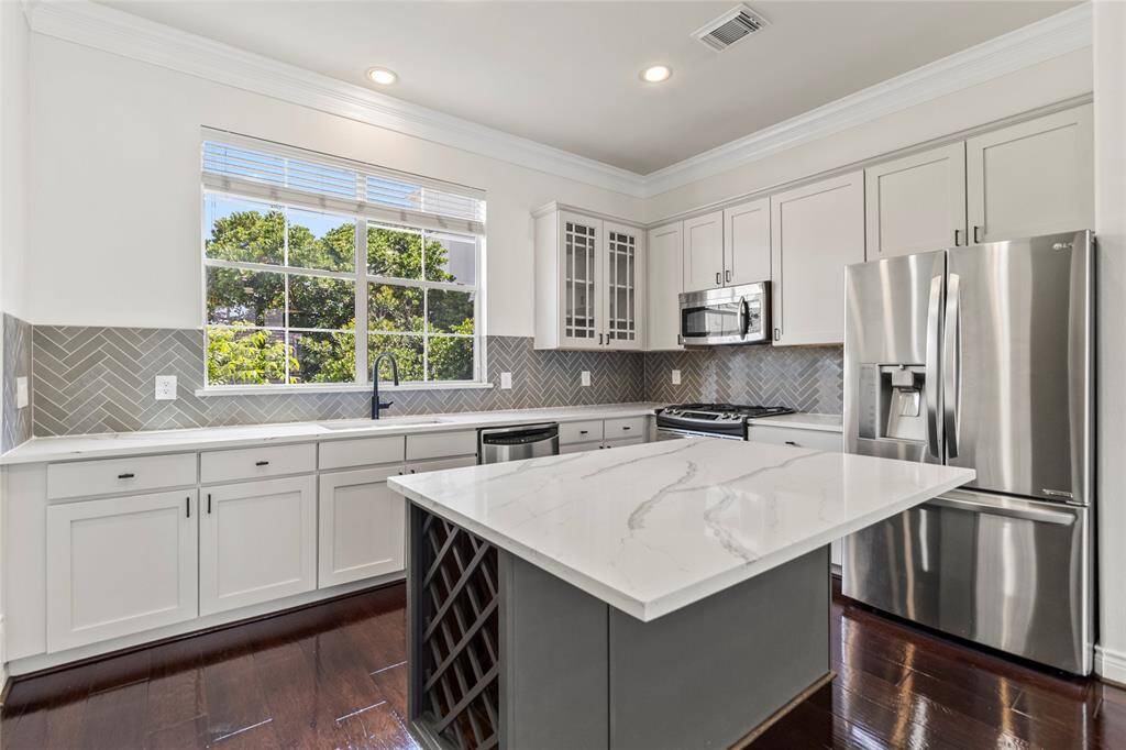 Crisp white cabinetry, quartz countertops, and a modern herringbone backsplash give your kitchen a bright, clean feel. Stainless steel appliances and a deep undermount sink enhance both function and style, while the oversized island with built-in wine storage adds a designer touch. Natural light streams through the large window, making the whole space feel airy and inviting.