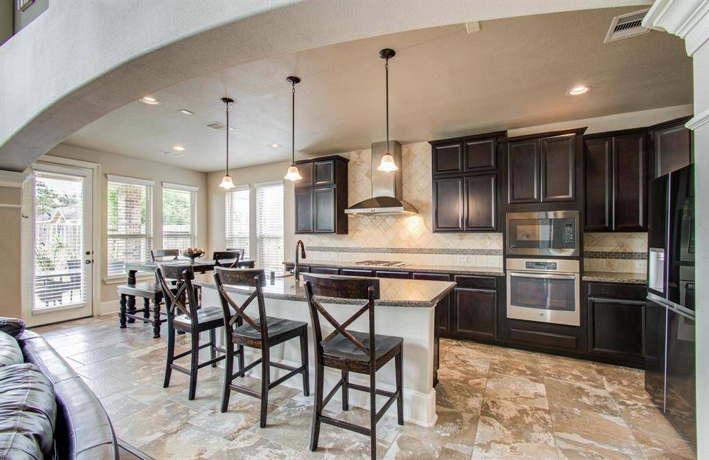 Modern kitchen with dark cabinetry, stainless steel appliances, and a central island with seating. The space is well-lit with pendant lights and large windows, featuring an open layout that connects to a dining area.