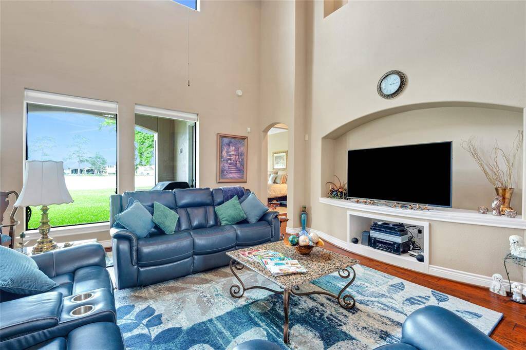Spacious family room with high ceilings and abundant natural light. Features include a large TV niche, elegant decor, and comfortable seating. Windows offer a picturesque view of the lake.
