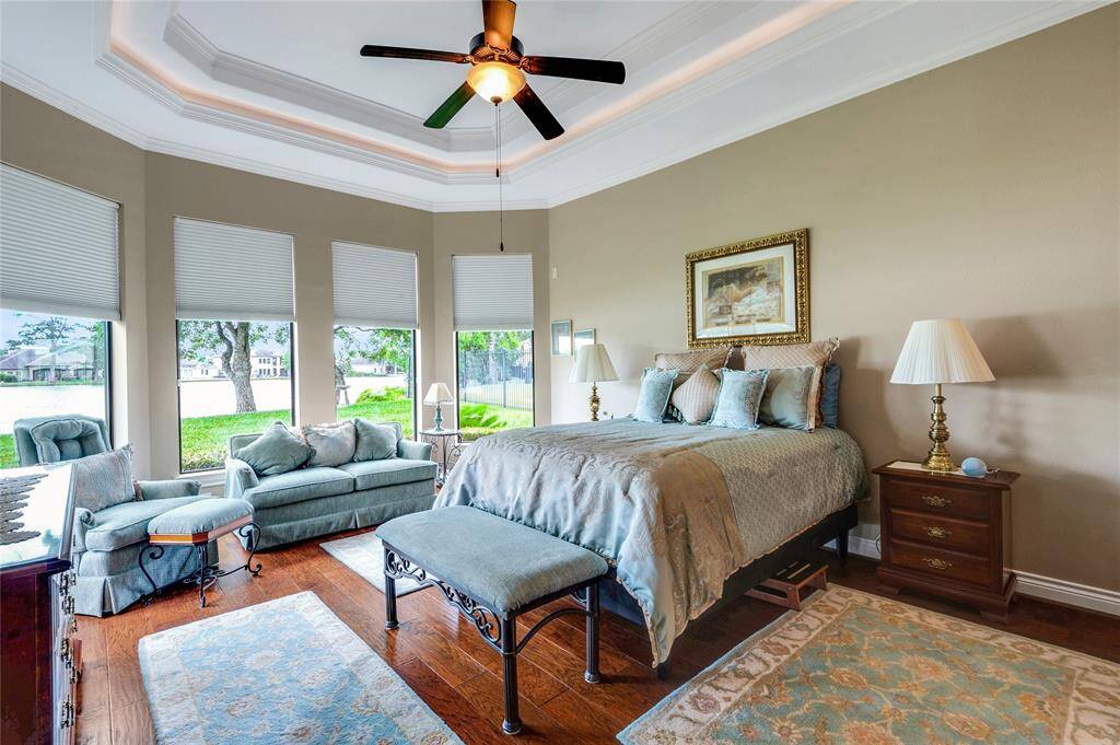 Bright and spacious bedroom with large windows offering lake views. Features a cozy sitting area, elegant tray ceiling with fan, and hardwood floors. Perfect for relaxation and comfort.