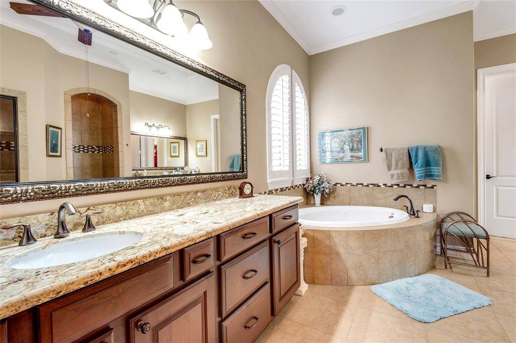 This bathroom features a spacious layout with a luxurious corner tub, dual sinks set in a granite countertop, elegant cabinetry, and a oversize designer mirror. The neutral tones and arch window light create a relaxing atmosphere.