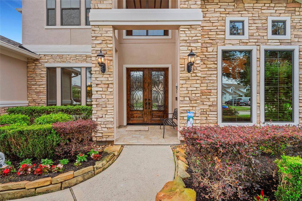 This charming home entry features a stone and stucco facade, elegant double doors with decorative glass, and well-maintained landscaping. Large windows provide natural light, and the inviting pathway leads to a cozy sitting area.