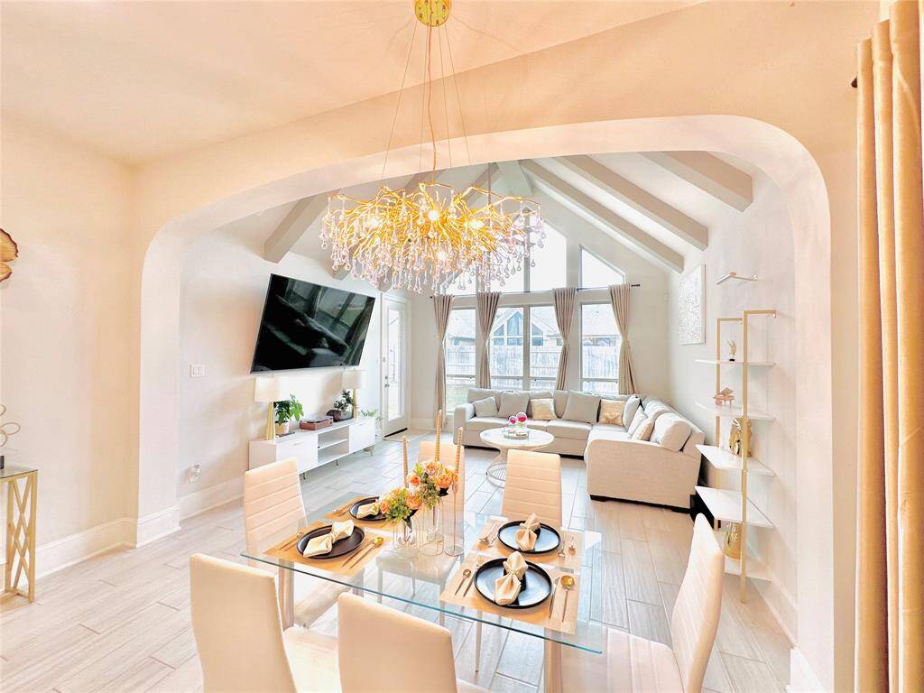 Open-plan living and dining space with high ceilings, large windows allowing ample natural light, an elegant chandelier, and contemporary furnishings. The room features neutral tones, creating a warm and inviting atmosphere.
