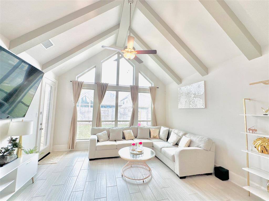 Living room featuring high vaulted ceilings with beams, large windows allowing for ample natural light, and a contemporary design with a neutral color palette. The room is furnished with a cozy sectional sofa and has stylish decor accents. A ceiling fan provides comfort, and the space opens to a backyard view through glass doors.
