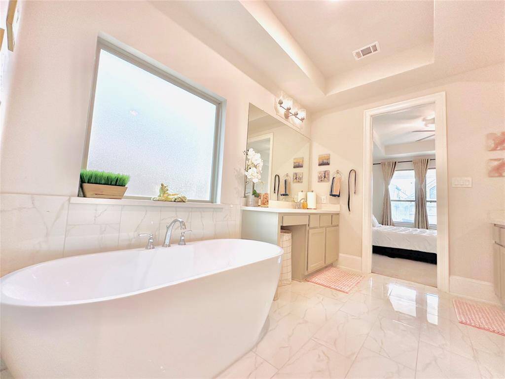 Bathroom featuring a standalone tub, a frosted window for privacy, dual vanity with ample counter space, and tiled flooring. A view of an adjoining room suggests a master suite layout.