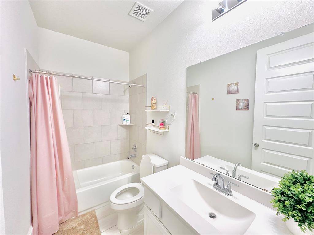 Well-lit, clean bathroom with a single vanity, a large mirror, a bathtub with a pink shower curtain, a toilet, and white cabinetry. It features neutral colors and modern fixtures, creating a serene and functional space.