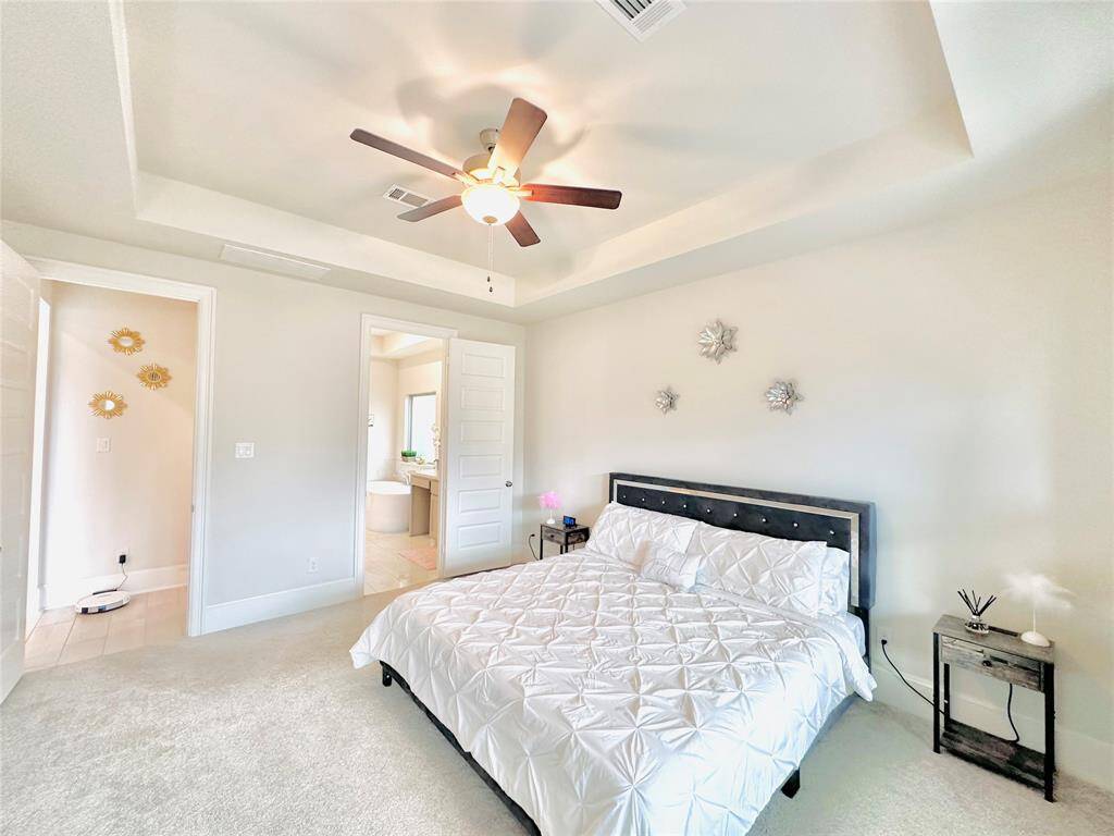 Primary bedroom featuring a vaulted ceiling with a ceiling fan, plush carpeting, and an en-suite bathroom. Decorative elements include wall art and a modern bedframe, with natural light streaming in from a window.