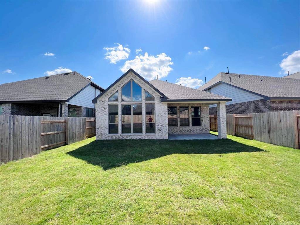 This is a sunny backyard view of a modern single-story brick house featuring large windows and a spacious lawn, enclosed by a wooden privacy fence.