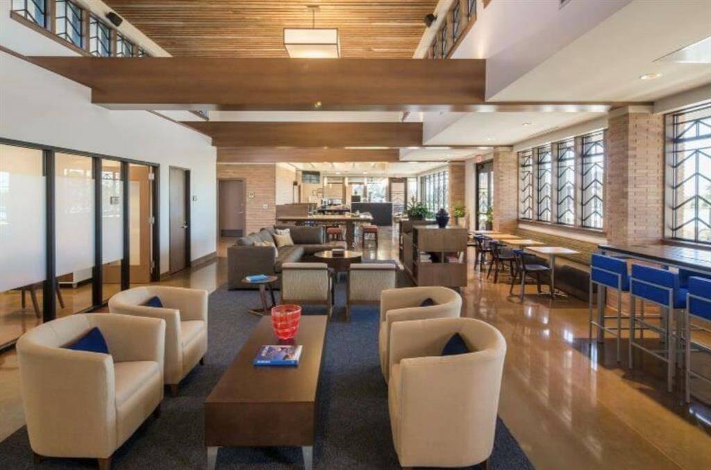 Open-plan living area with high ceilings, exposed beams, and large windows that allow for plenty of natural light. The room features a comfortable seating area with armchairs and a sofa, a dining area with stylish bar stools, and sleek, contemporary decor throughout.