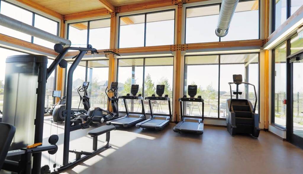 Gym with large windows allowing for plenty of natural light, featuring an array of cardio equipment and a weight training station. The room boasts high ceilings and a pleasant view, ideal for an energizing workout environment.