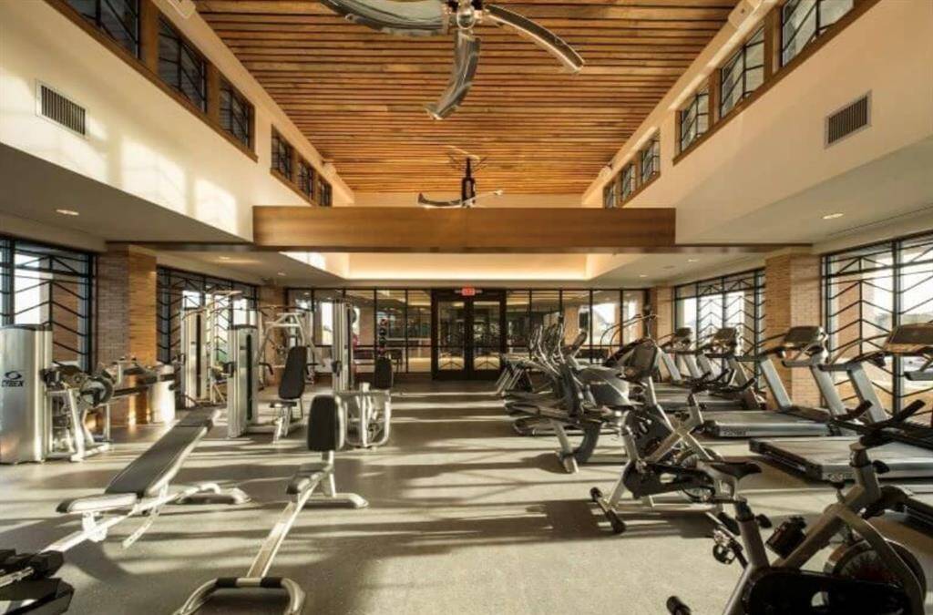 Well-equipped home gym with high ceilings, ample natural light, and a variety of exercise machines. The room features a wood-paneled ceiling and large windows, creating an inviting atmosphere for workouts.