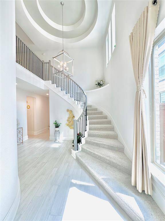 Bright and airy entryway featuring a sweeping staircase with a stylish metal railing, high ceilings with a modern pendant light, and large windows allowing plenty of natural light. The space has a fresh, contemporary feel with neutral colors and elegant flooring.
