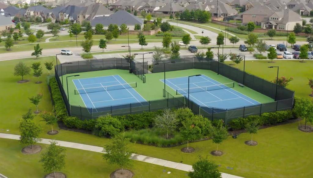 Tennis courts with blue playing surfaces and green outskirts, enclosed by a tall fence. The courts are part of a larger residential community with manicured green spaces and a variety of houses in the background. There are roads with some traffic adjacent to the courts, suggesting easy access.