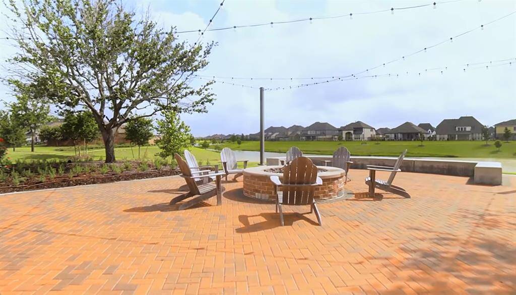 Outdoor patio with herringbone brick pavers, featuring a fire pit and comfortable seating under string lights, with a scenic view of a well-manicured community lawn and neighboring homes in the background.