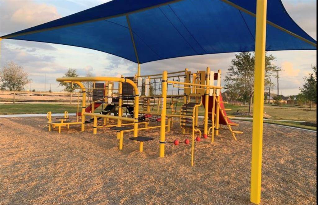 Community playground featuring a colorful play structure with slides and climbing elements, shaded by a large blue canopy, set in a park-like setting with open spaces in the background.