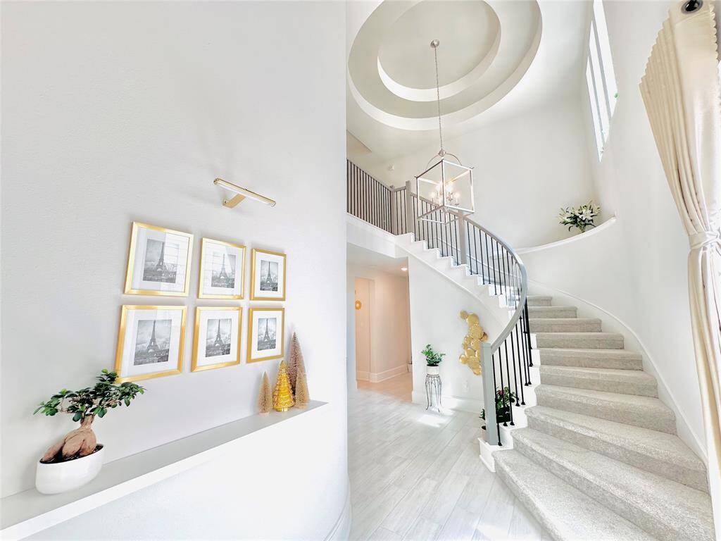 Spacious entryway featuring a grand staircase with a stylish wrought iron railing, leading up to a well-lit second floor. The foyer is accented with an elegant chandelier, high ceilings, and a tasteful arrangement of framed pictures on the wall. The neutral color palette and the natural light streaming through the windows create a welcoming atmosphere.