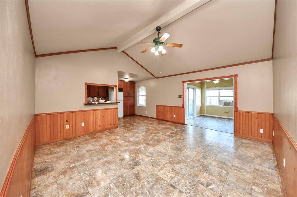 This open-concept living area features soaring vaulted ceilings with exposed beams, adding a sense of space and character. The natural wood trim and wainscoting give the room a warm, inviting feel, while the neutral tile flooring is both stylish and easy to maintain. A pass-through window to the kitchen enhances functionality and connection. Large entry leads into a bright sunroom, perfect for relaxing or entertaining.