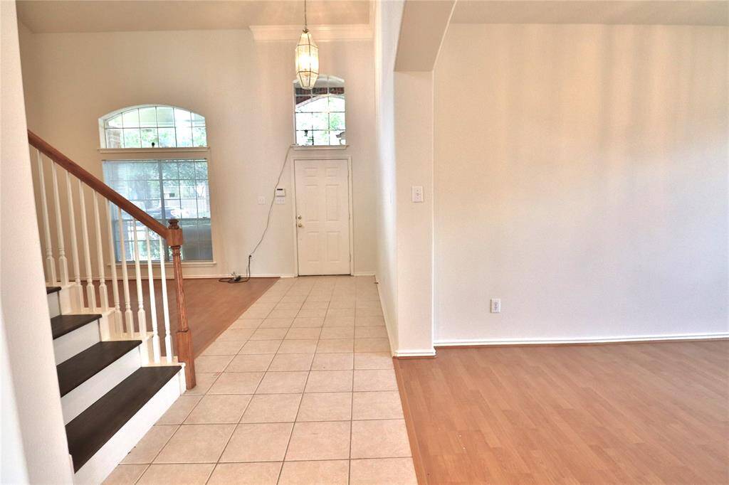 This entryway features a tiled foyer leading to a wooden staircase. It has high ceilings,  and ample natural light from large windows, offering a welcoming and airy atmosphere.
