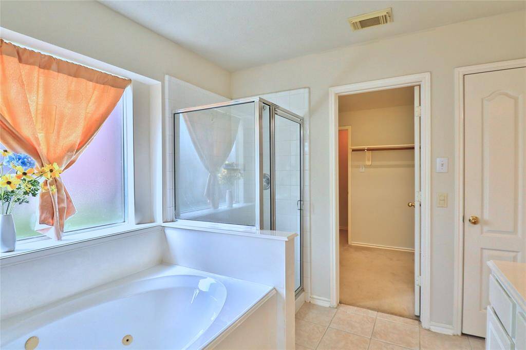 This primary bathroom features a large soaking tub and a separate glass-enclosed shower. A window with curtains allows natural light, and there's an adjoining walk-in closet for convenience.