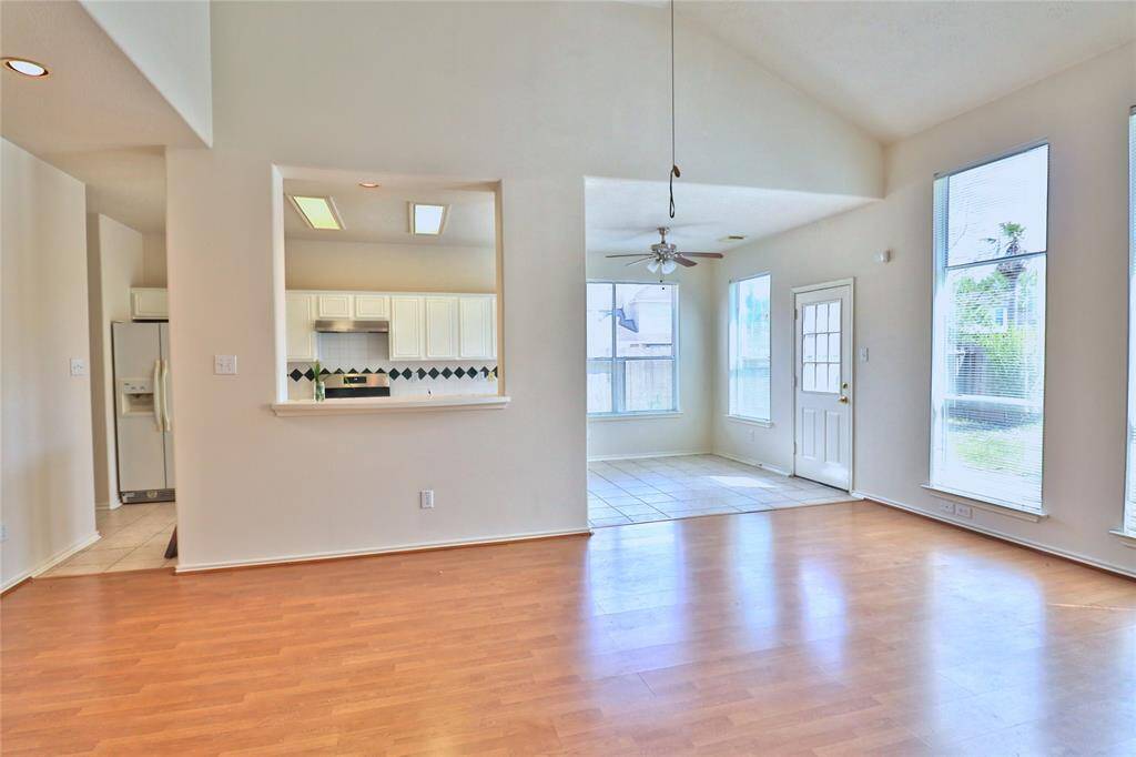 This bright, open-concept space features large windows for natural light, and a view into the kitchen with white cabinetry. The adjacent dining area has tiled flooring and a ceiling fan, leading to a door opening to the backyard.