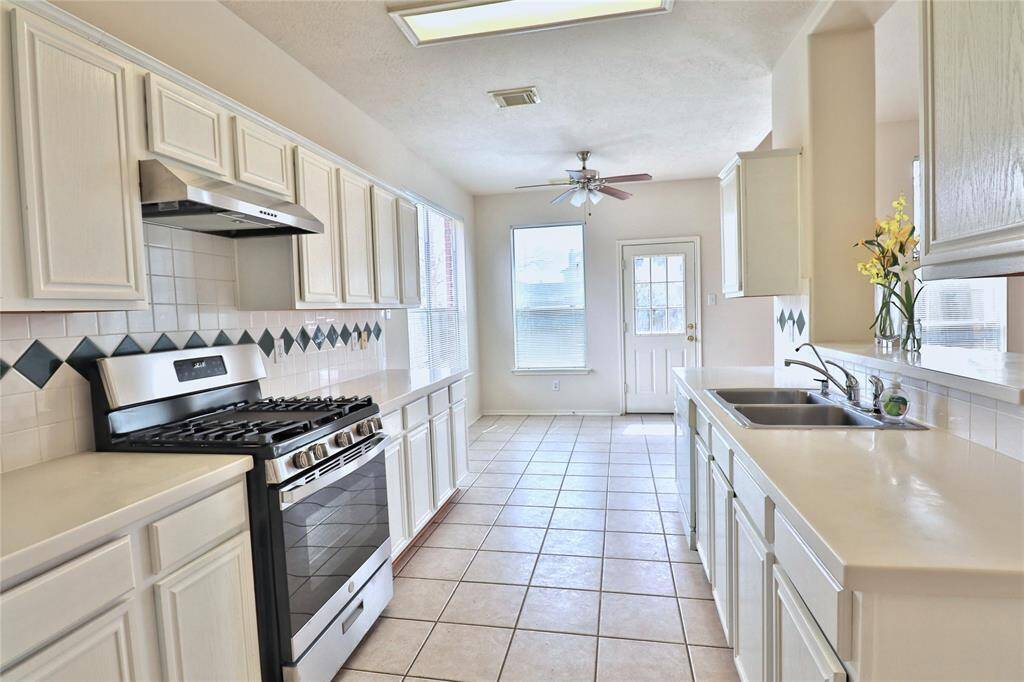 Bright, spacious kitchen with white cabinetry and tile flooring. Features a modern gas stove, ample counter space, and dual sinks. Natural light fills the room through large windows and a glass-paneled door, leading to an inviting atmosphere.