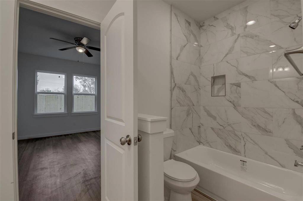 Experience modern luxury in this bright bathroom featuring a marble-tiled shower and bathtub, seamlessly connected to a bedroom with sleek vinyl wood-style flooring and a ceiling fan. Large windows fill the space with natural light.
