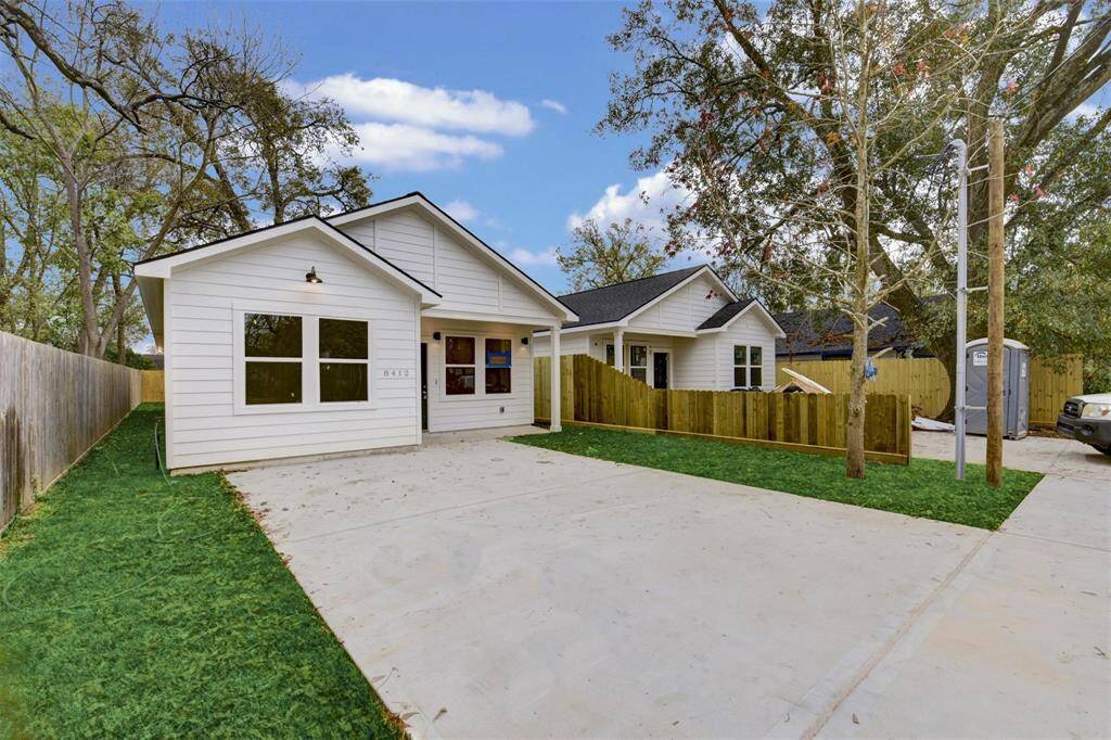 This photo shows a modern, single-story white house with a gable roof. The property features a large driveway, fenced yard, and a small front lawn with trees, offering a clean and inviting curb appeal.