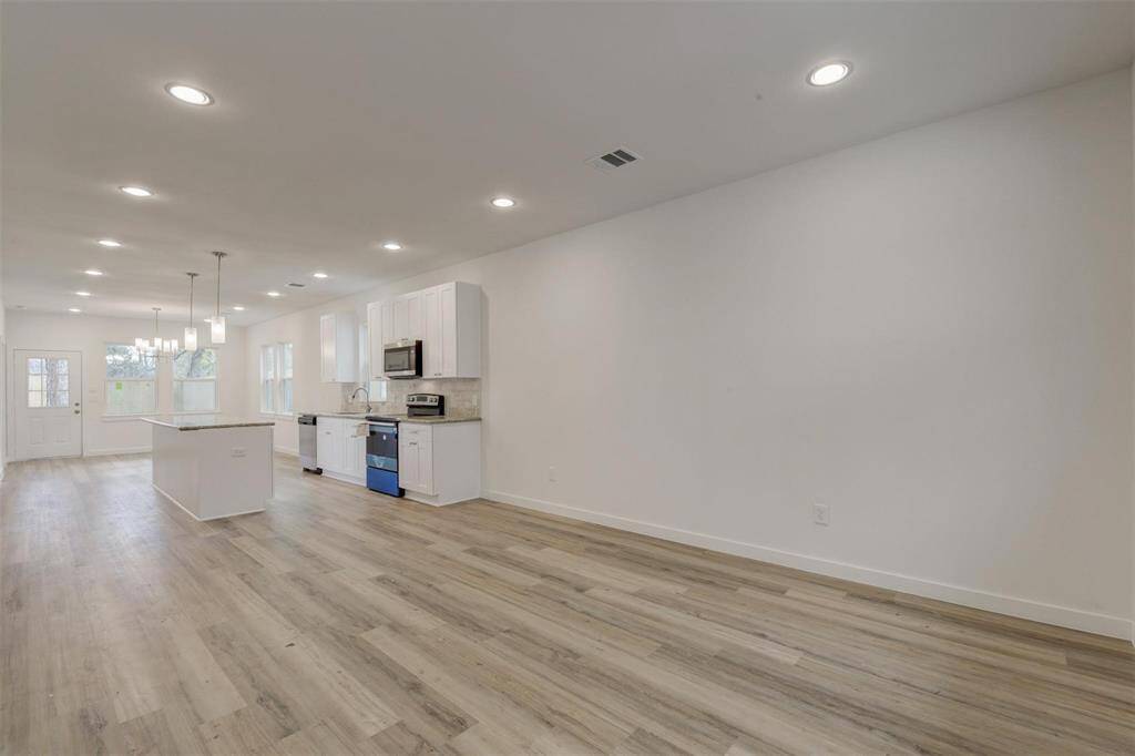 This photo shows a spacious, open-concept living area with modern kitchen features, including white cabinets, stainless steel appliances, and a central island. The room is well-lit with recessed lighting and has light vinyl wood flooring, creating a bright and inviting atmosphere.