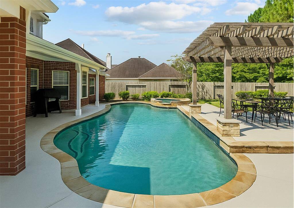 This backyard features a sleek in-ground pool with a stone border, a cozy hot tub, and a spacious patio area with a pergola for outdoor dining and relaxation. The surrounding privacy fence and lush greenery create a serene and private oasis.