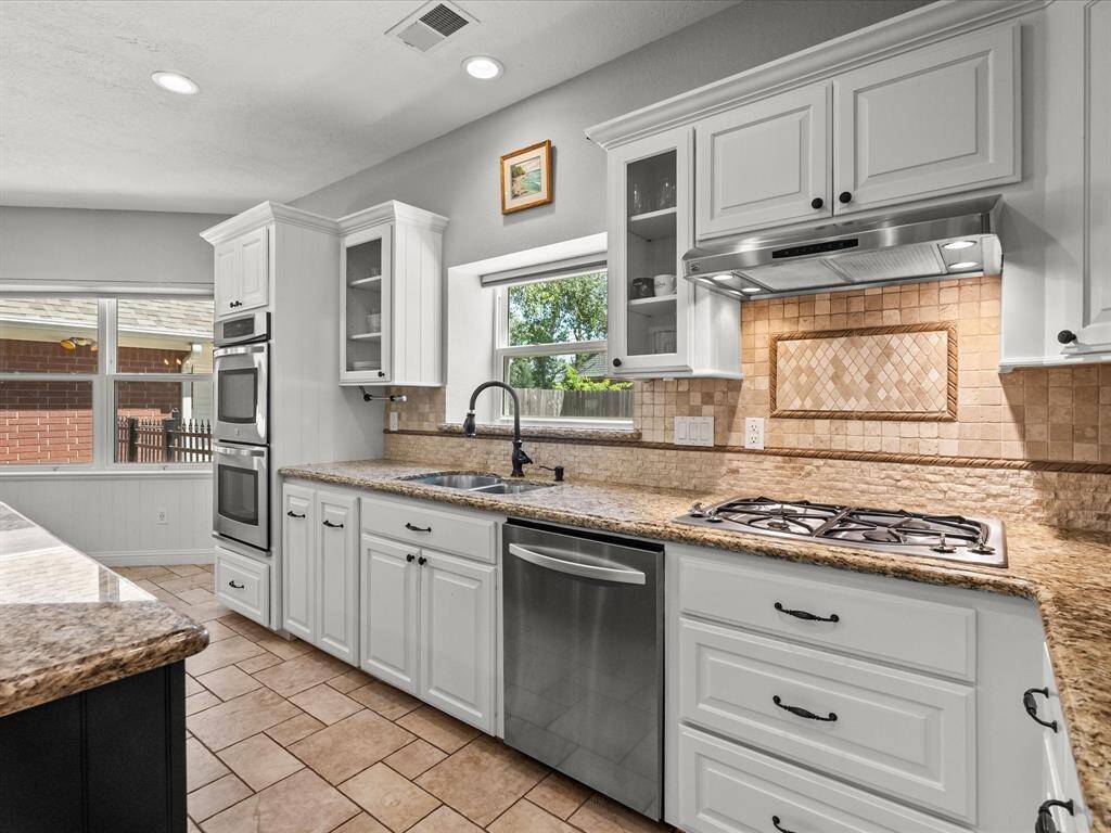 The kitchen features elegant white cabinetry with glass accents, granite countertops, and a tile backsplash. It includes a built-in double oven, gas cooktop, and stainless steel dishwasher, offering a bright and functional cooking space.
