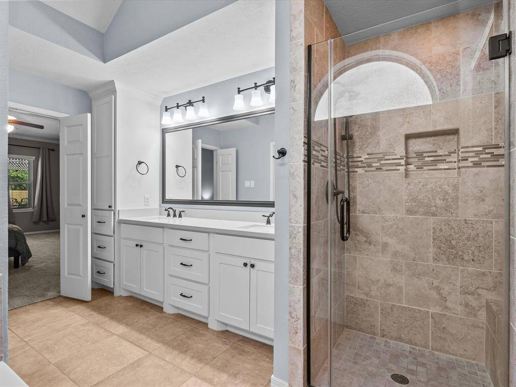This photo showcases a modern bathroom with a spacious tiled shower, glass door, and intricate tile accents. The vanity features dual sinks, ample storage, and elegant lighting.