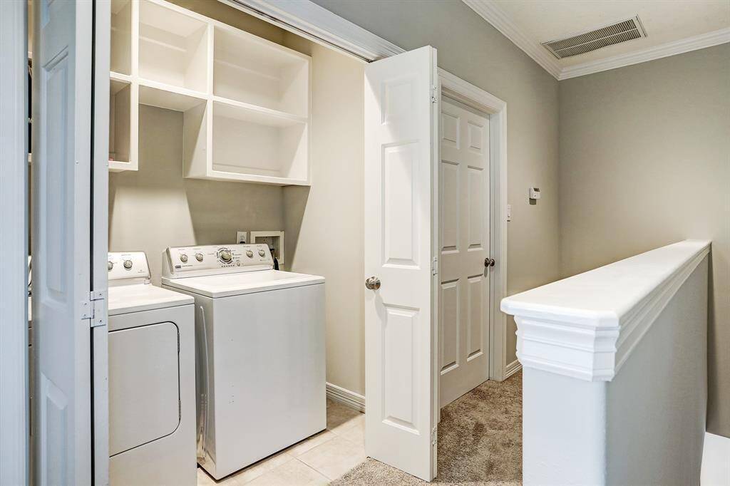 This photo showcases a compactlaundry area with a washer and dryer,open shelving, and neutral wall colors.It's conveniently located next to ahallway with carpet flooring and whitetrim.
