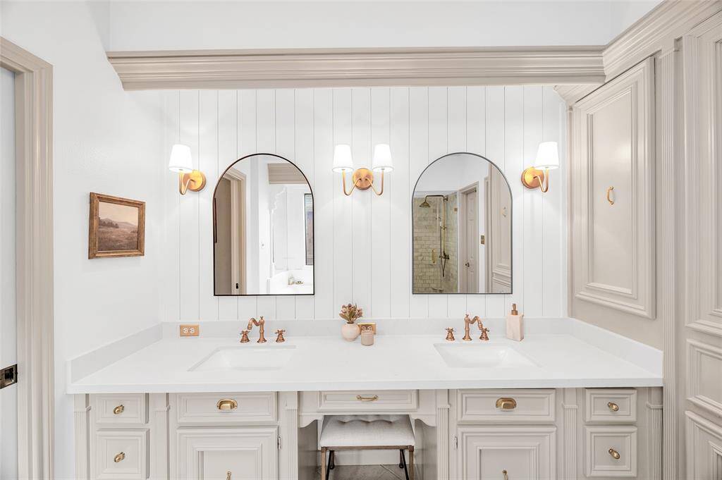 This bathroom features a double vanity with elegant white countertops and brass fixtures. Two arched mirrors are flanked by stylish wall sconces, and the cabinetry offers ample storage space. The neutral color palette and shiplap accent wall add a touch of modern sophistication.