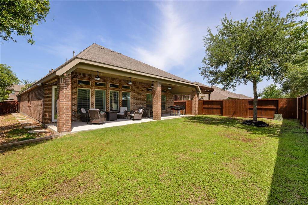 This photo showcases a spacious backyard with a covered patio attached to a brick house. The area features outdoor seating and a small dining space, surrounded by a wooden fence and a well-maintained lawn, perfect for relaxation and entertaining.