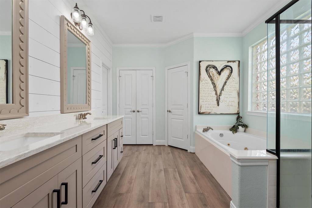 Bright and modern bathroom featuring dual sinks with large mirrors, a spacious bathtub and a glass-enclosed shower. The room is accented with light wood flooring and soft-toned walls for a spa-like feel.