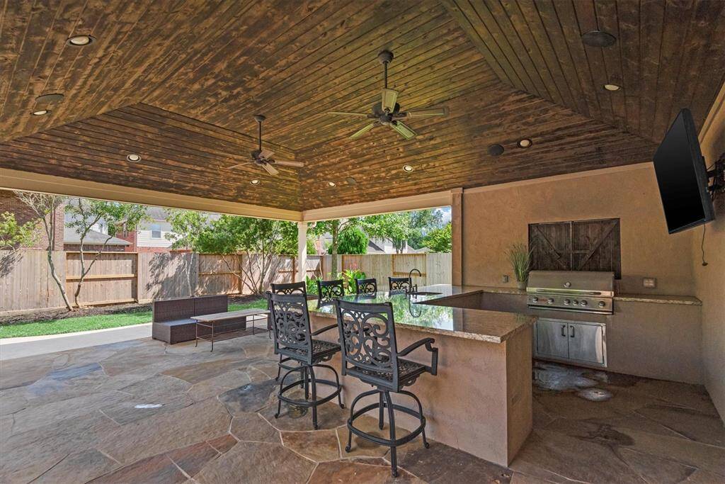 Large covered patio with a wooden ceiling, ceiling fans, and recessed lighting. It includes a built-in grill, sink and bar seating.